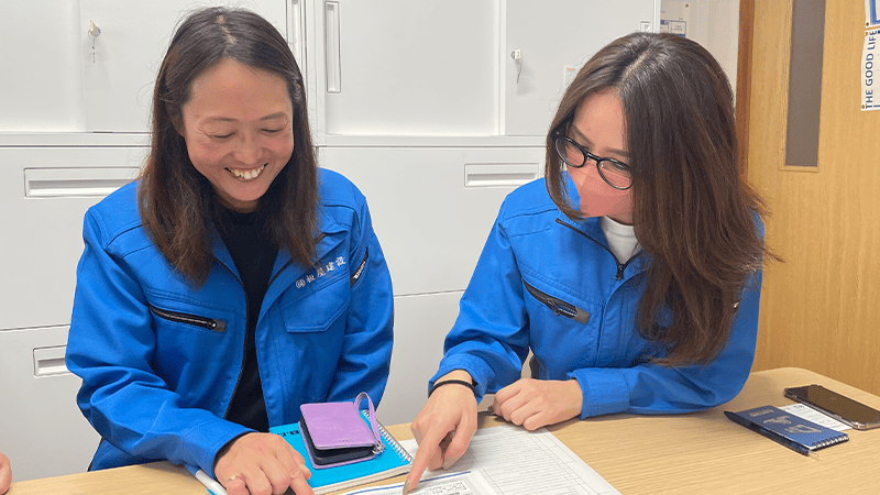 オフィスで作業着の女性が二人並んで資料を指さしている様子