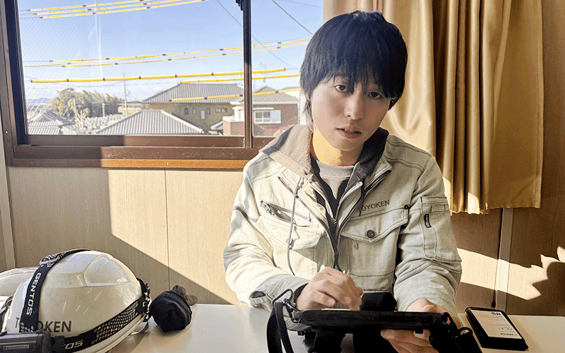 屋内でタブレットを操作している作業着の男性の写真。