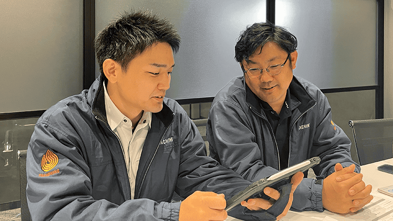 屋内でiPadを見ながら会話する二人の作業服の男性の写真